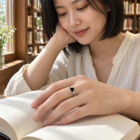 A sterling silver ring with a black center stone is worn on a hand resting across an open blank book near a wooden window.