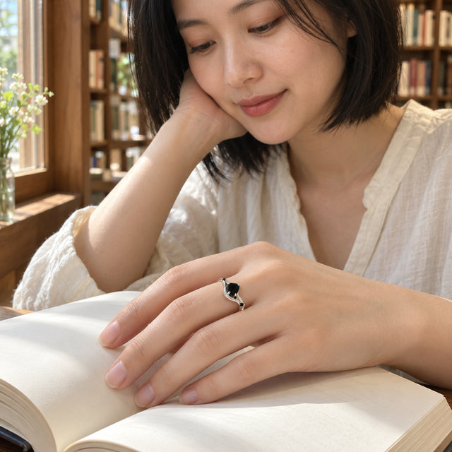 A sterling silver ring with a black center stone is worn on a hand resting across an open blank book near a wooden window.