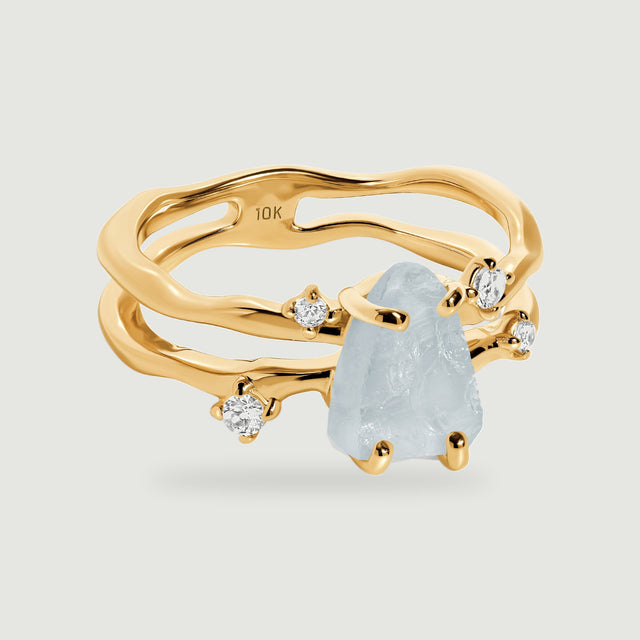 A yellow gold ring with a pale blue raw aquamarine and four clear accent stones rests on a light gray surface.