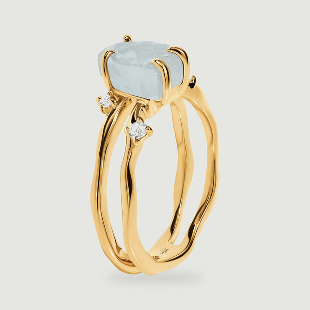 The yellow gold ring is shown from the side with an elevated pale blue crystal and two curved bands.