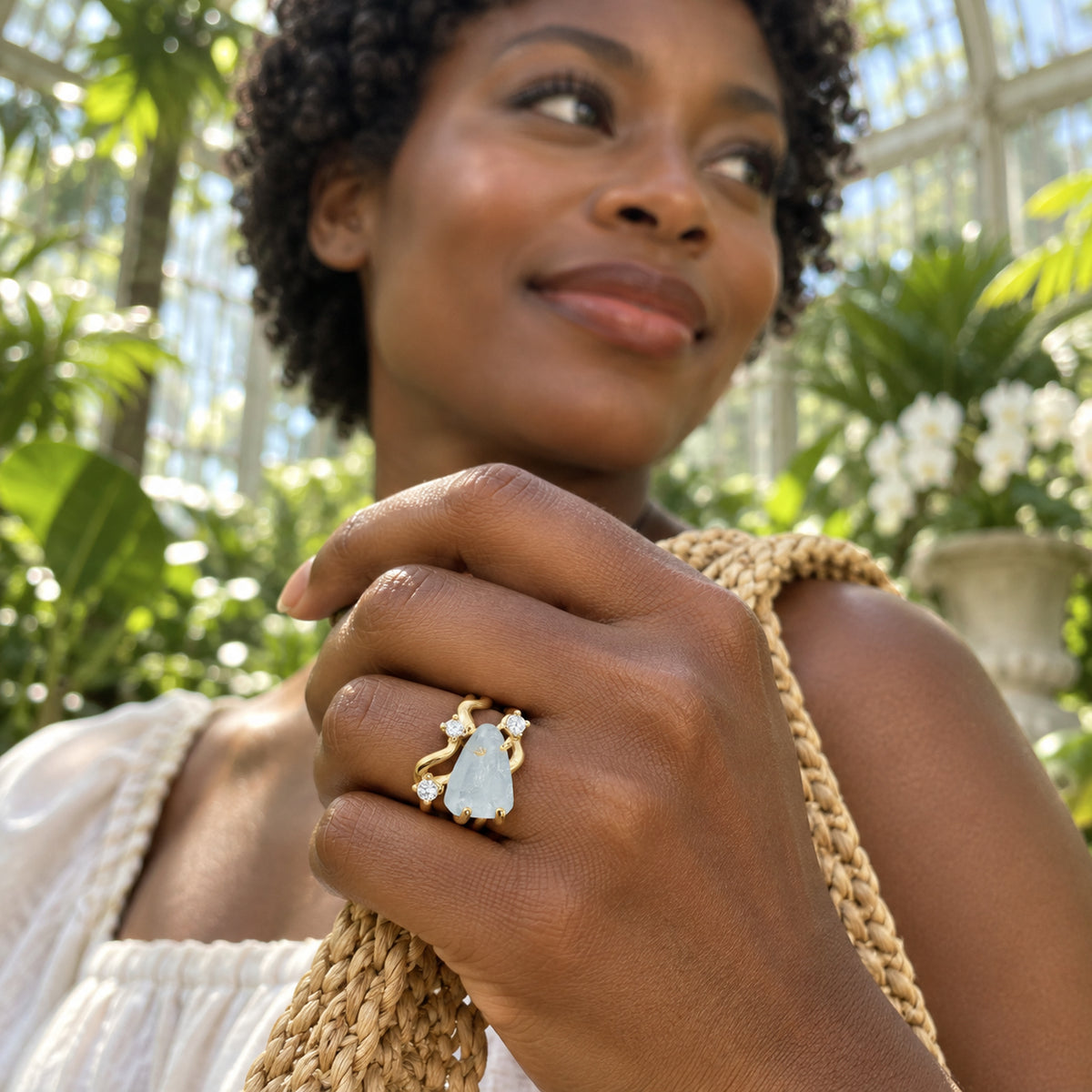 A yellow gold aquamarine ring is worn on a hand holding a woven bag strap among sunlit greenhouse plants.