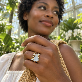 A yellow gold aquamarine ring is worn on a hand holding a woven bag strap among sunlit greenhouse plants.