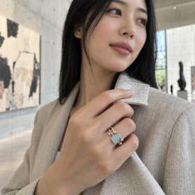 A white gold aquamarine ring is worn on a hand adjusting a linen jacket collar in an art gallery.