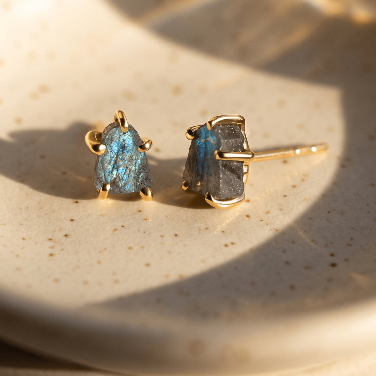 Two yellow gold-colored labradorite studs rest on a speckled ceramic dish with sunlight falling across the blue-gray stones.