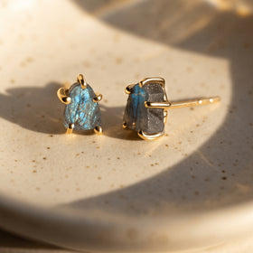 Two yellow gold-colored labradorite studs rest on a speckled ceramic dish with sunlight falling across the blue-gray stones.