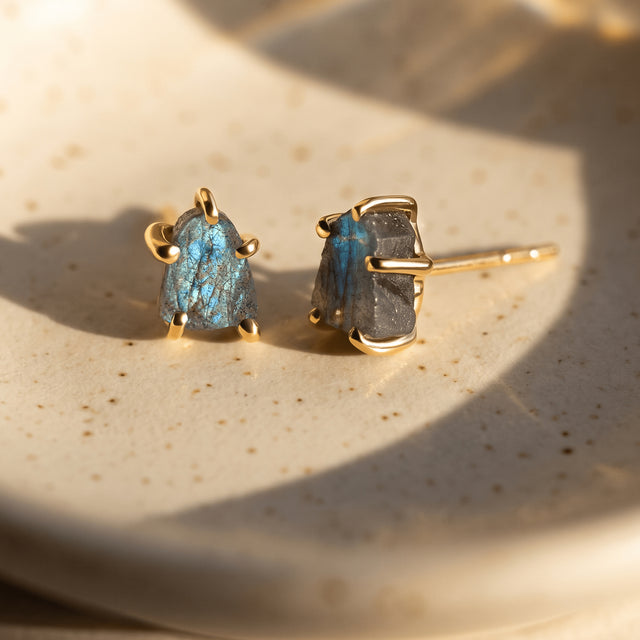 Two yellow gold-colored labradorite studs rest on a speckled ceramic dish with sunlight falling across the blue-gray stones.