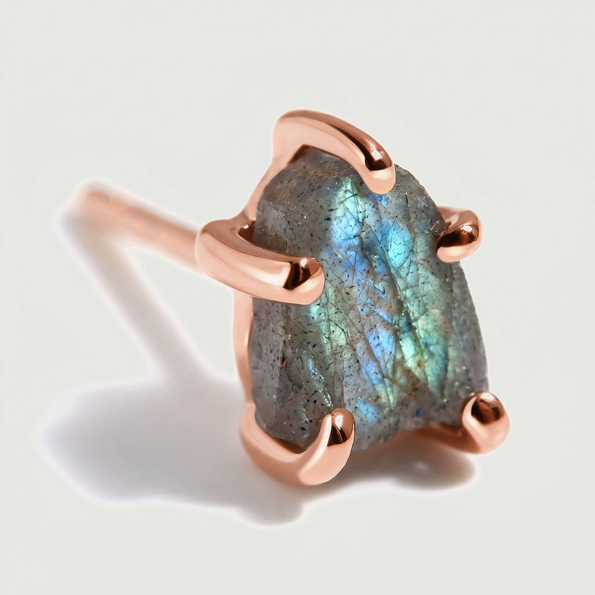 A raw labradorite stud shows blue and green flashes between polished rose gold-colored prongs, with its post softly blurred behind it.