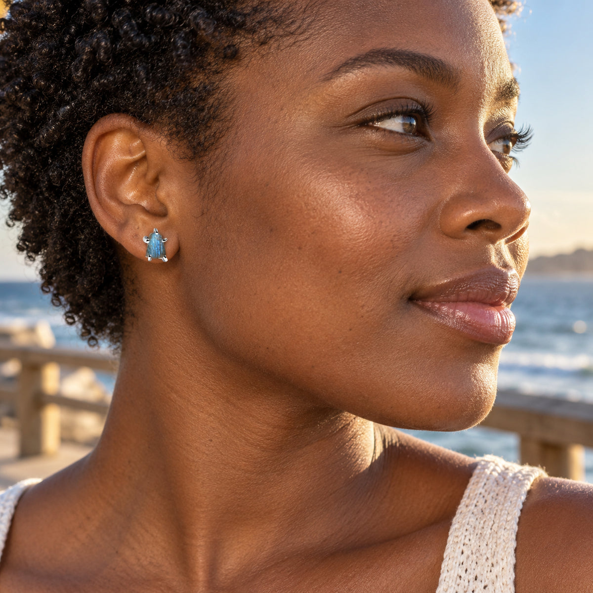 A woman with short coiled hair wears a blue labradorite stud in silver-colored prongs beside a sunlit seaside boardwalk.