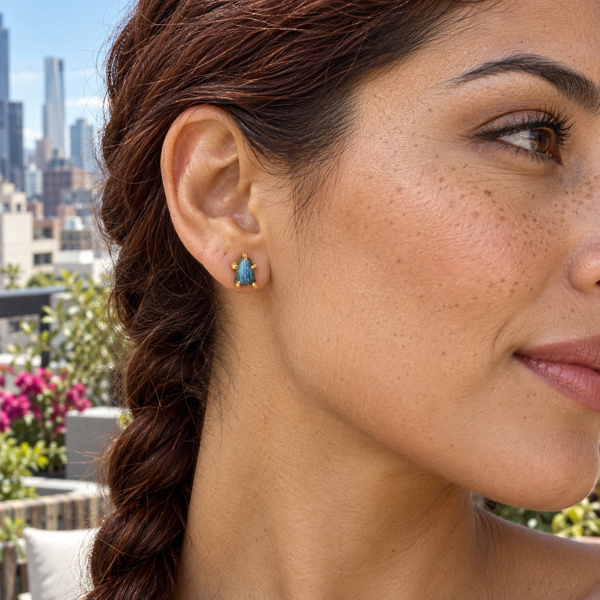 A woman with an auburn braid wears a raw blue labradorite stud in yellow gold-colored prongs on a rooftop terrace with buildings behind her.