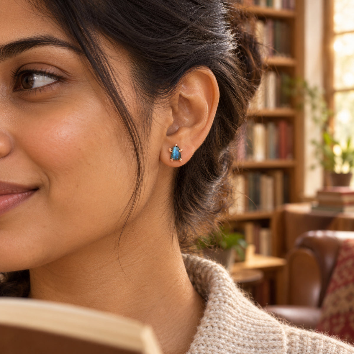 A woman with dark hair in a loose bun wears a blue-gray labradorite stud with rose gold-colored prongs beside blurred bookshelves.