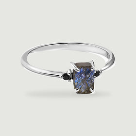 A sterling silver ring with a rectangular blue-grey labradorite and two black side stones rests on a light grey surface.