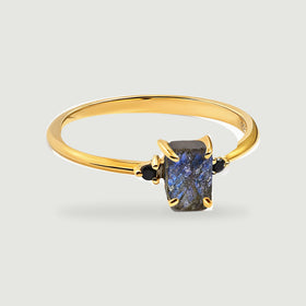 A yellow gold vermeil ring with a rectangular blue-grey labradorite and two black side stones rests on a light grey surface.