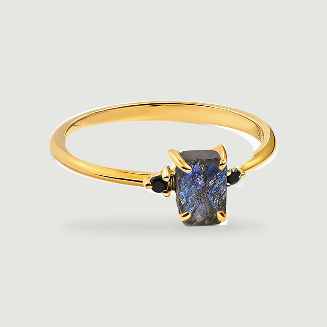 A yellow gold vermeil ring with a rectangular blue-grey labradorite and two black side stones rests on a light grey surface.