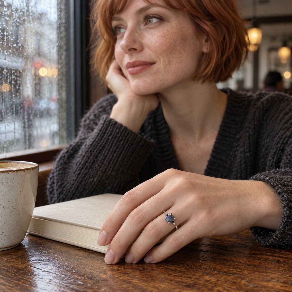 A woman with short copper hair wears the sterling silver ring while resting her hand on a closed book beside a rain-speckled window.