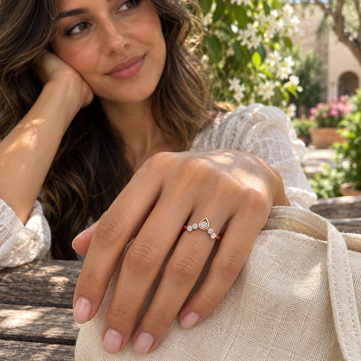 A woman wears a rose gold five-diamond ring while resting her hand on a linen bag in a flowering courtyard.