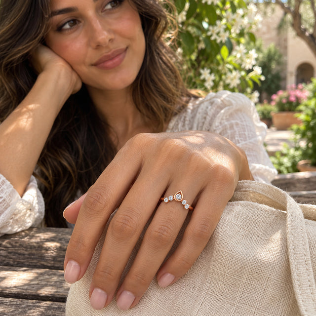 A woman wears a rose gold five-diamond ring while resting her hand on a linen bag in a flowering courtyard.