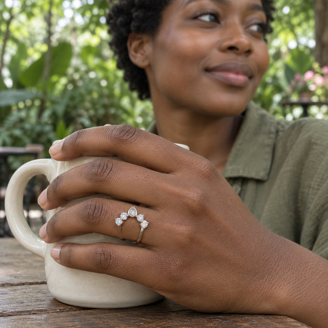 A woman wearing a white gold five-diamond ring holds an ivory ceramic mug at a leafy outdoor cafe.