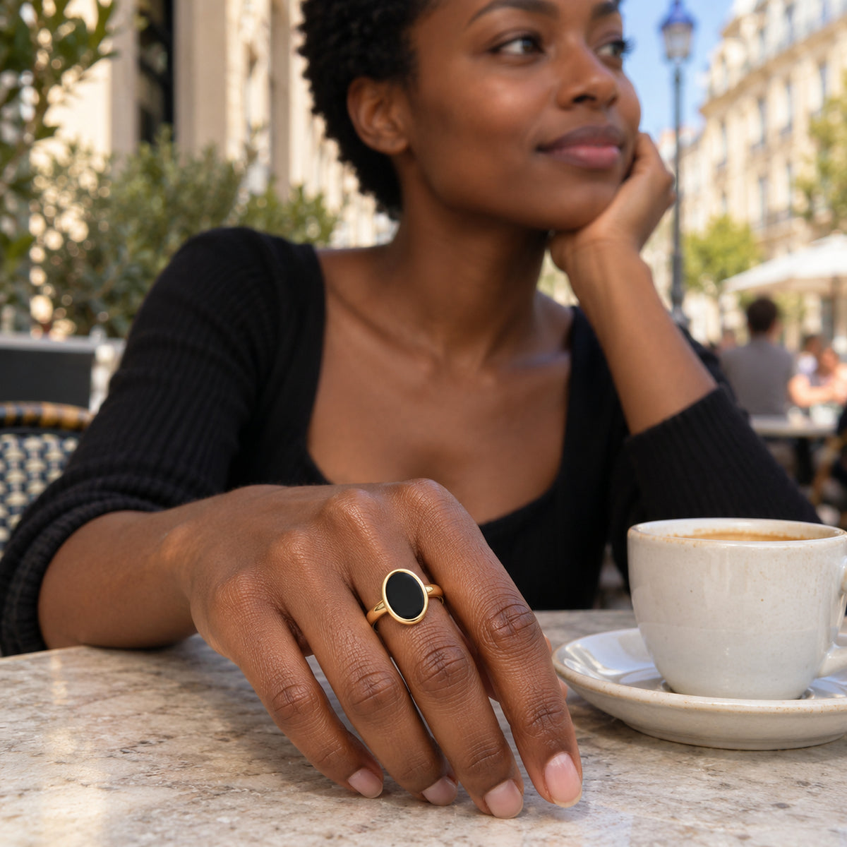 A yellow gold vermeil black onyx ring is worn on a hand beside a ceramic coffee cup at an outdoor cafe.