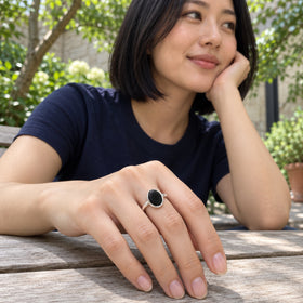 An oval black onyx ring in sterling silver is shown on a hand resting on a wooden bench beneath leafy trees.