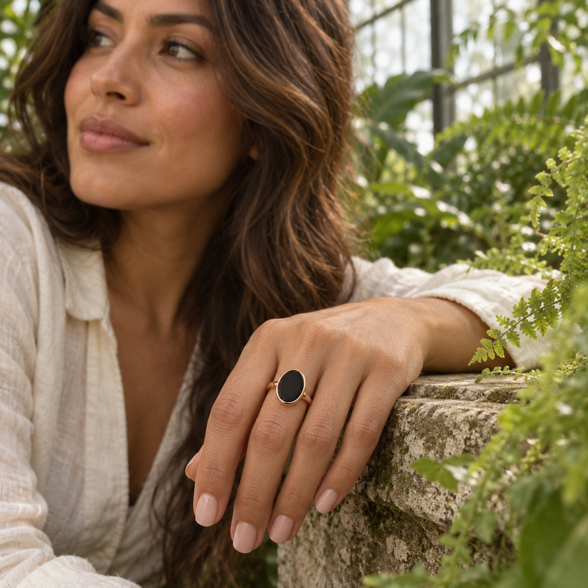 A rose gold vermeil black onyx ring is worn on a hand resting on a stone ledge among greenhouse ferns.