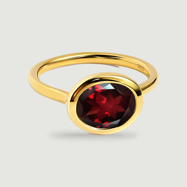 An oval red garnet sits in a polished yellow gold bezel on a slender ring, centered against a light gray background.