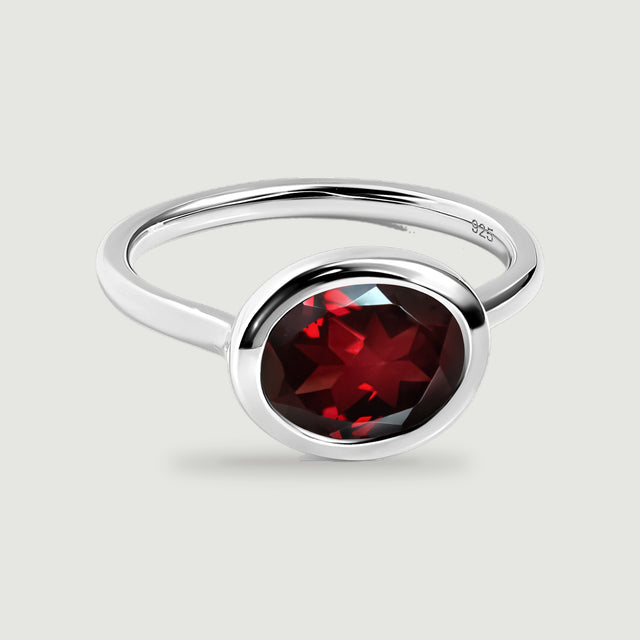 A silver ring with a deep red faceted garnet is shown at a three-quarter angle on pale gray.