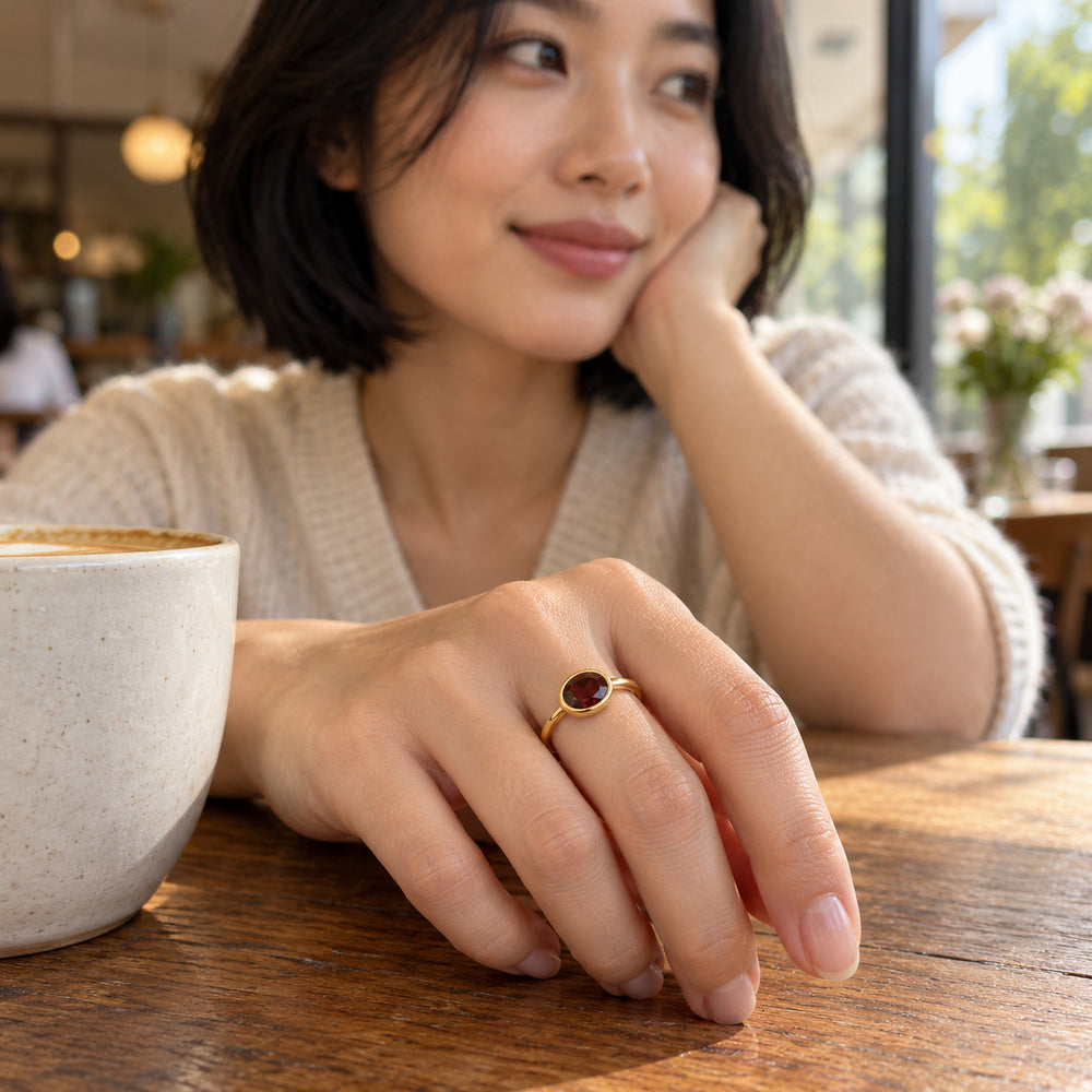 A yellow gold garnet ring is worn on a hand resting beside a ceramic cup on a wooden cafe table.