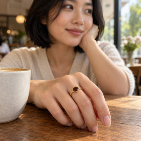 A yellow gold garnet ring is worn on a hand resting beside a ceramic cup on a wooden cafe table.