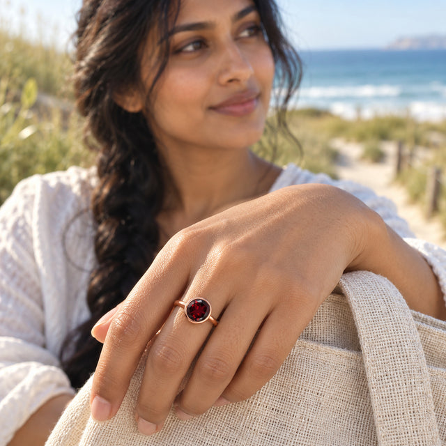 A rose gold garnet ring catches the light on a hand holding a linen tote near the coast.