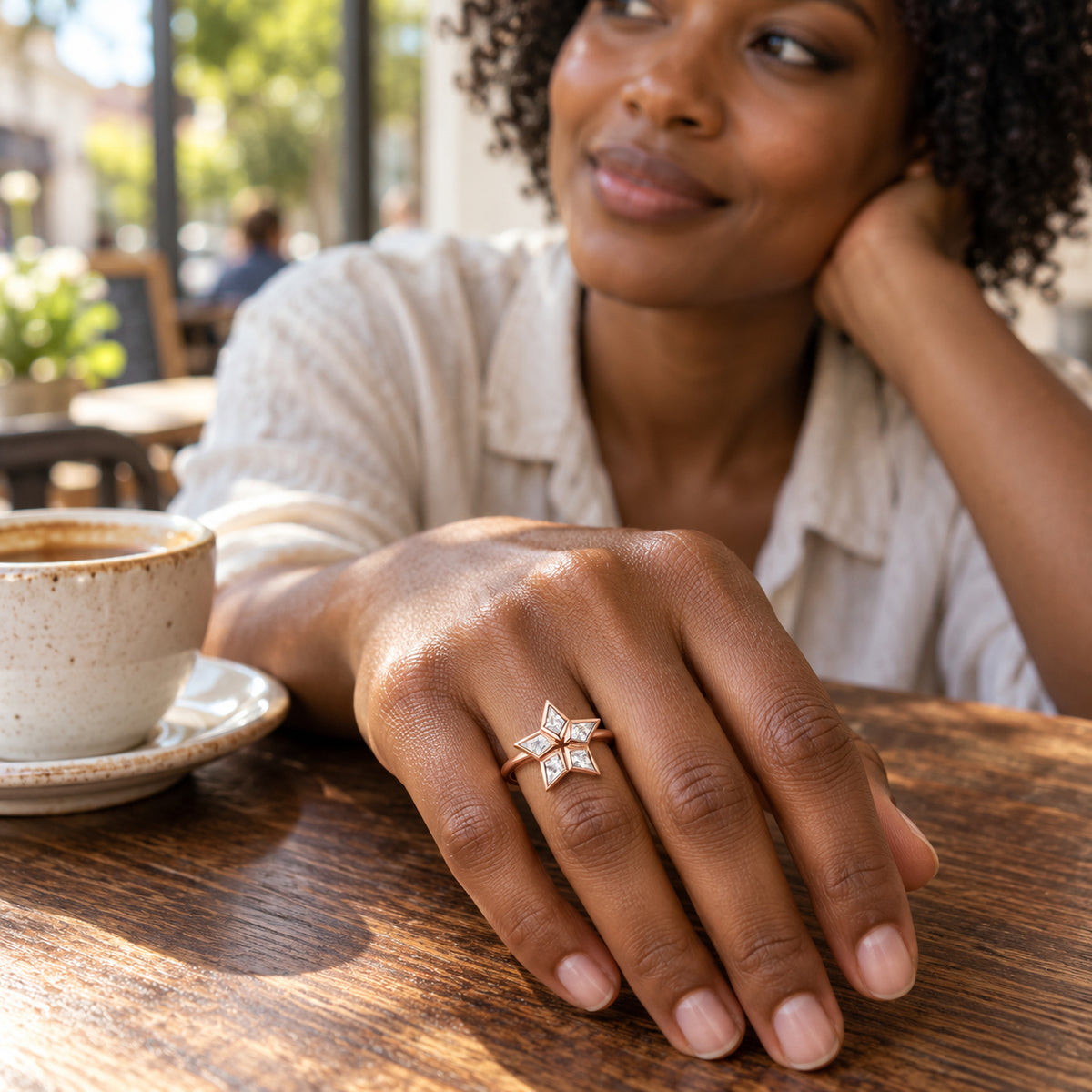 A rose gold star-shaped ring with five clear stones is worn on a hand resting beside a ceramic cup on a wooden cafe table.