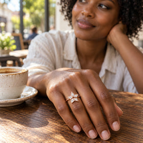 A rose gold star-shaped ring with five clear stones is worn on a hand resting beside a ceramic cup on a wooden cafe table.