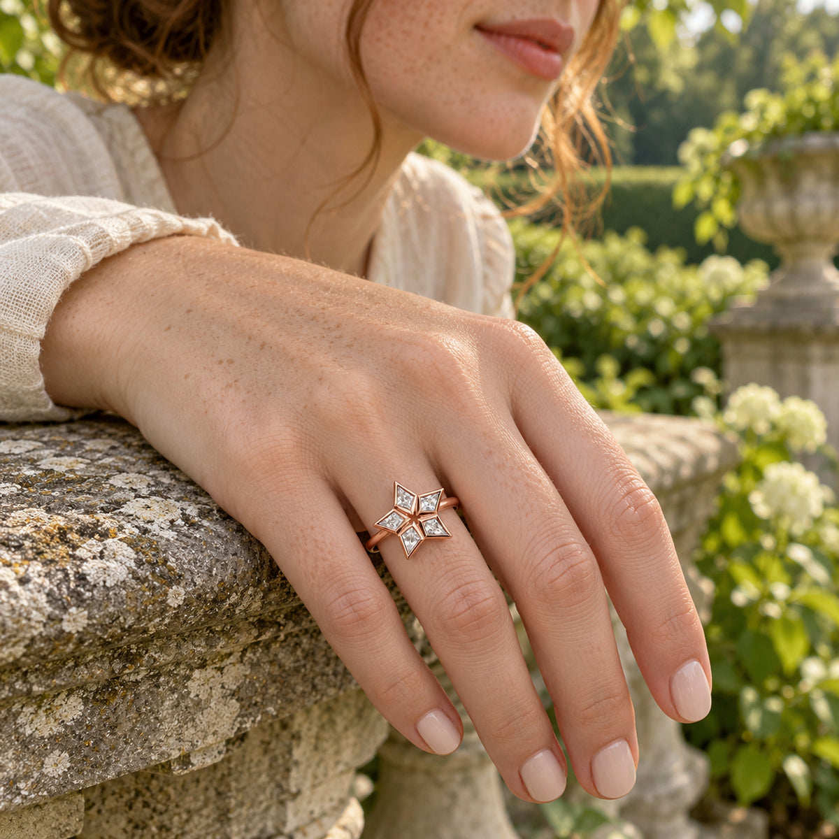 A five-stone rose gold ring is shown on a freckled hand resting on a weathered stone rail with green garden foliage behind it.