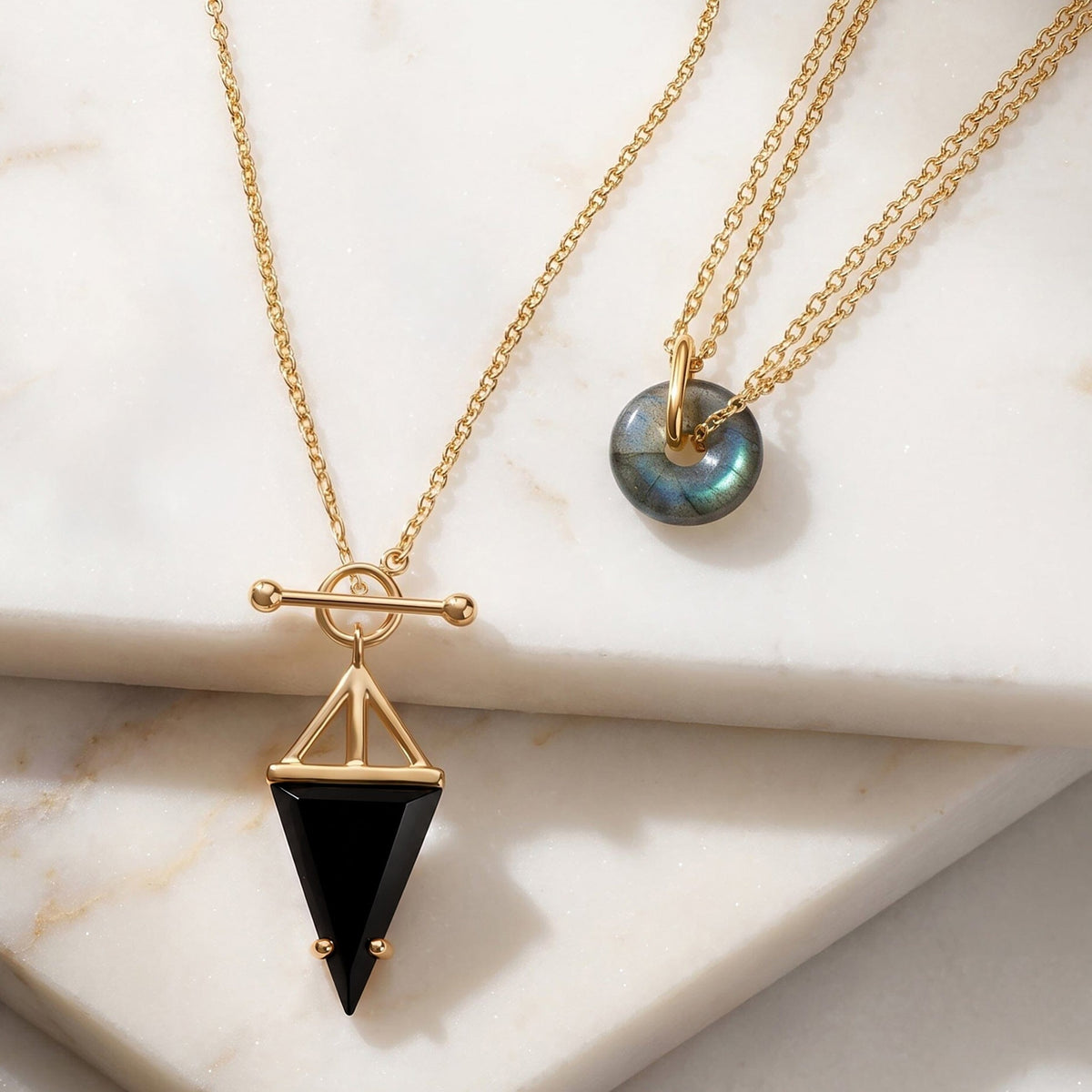 Gold necklaces, one with a labradorite donut and one with a black obsidian pendant, on a marble surface