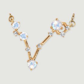 The complete V-shaped gold pendant lies against a light grey surface with both chain attachments visible.