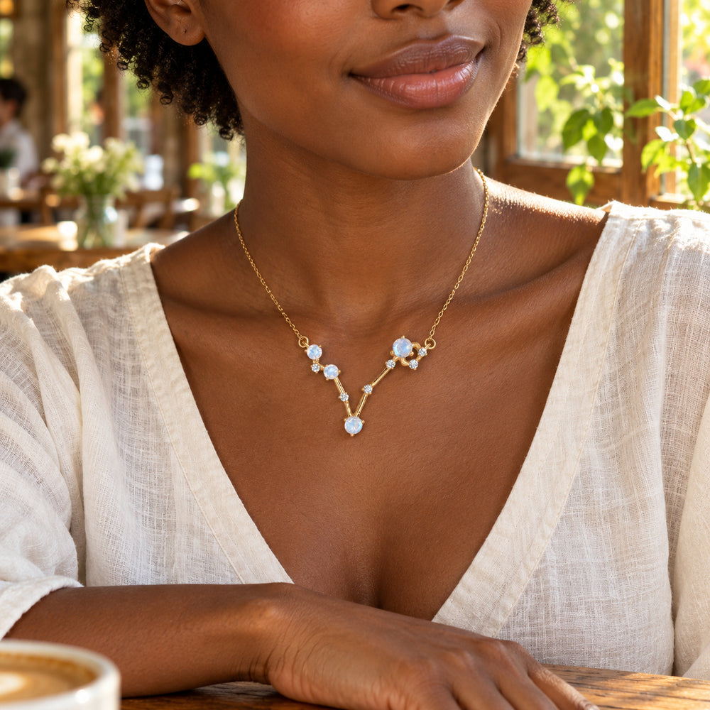 A woman with short curls wears the gold moonstone necklace with an ivory blouse beside a sunlit cafe window.