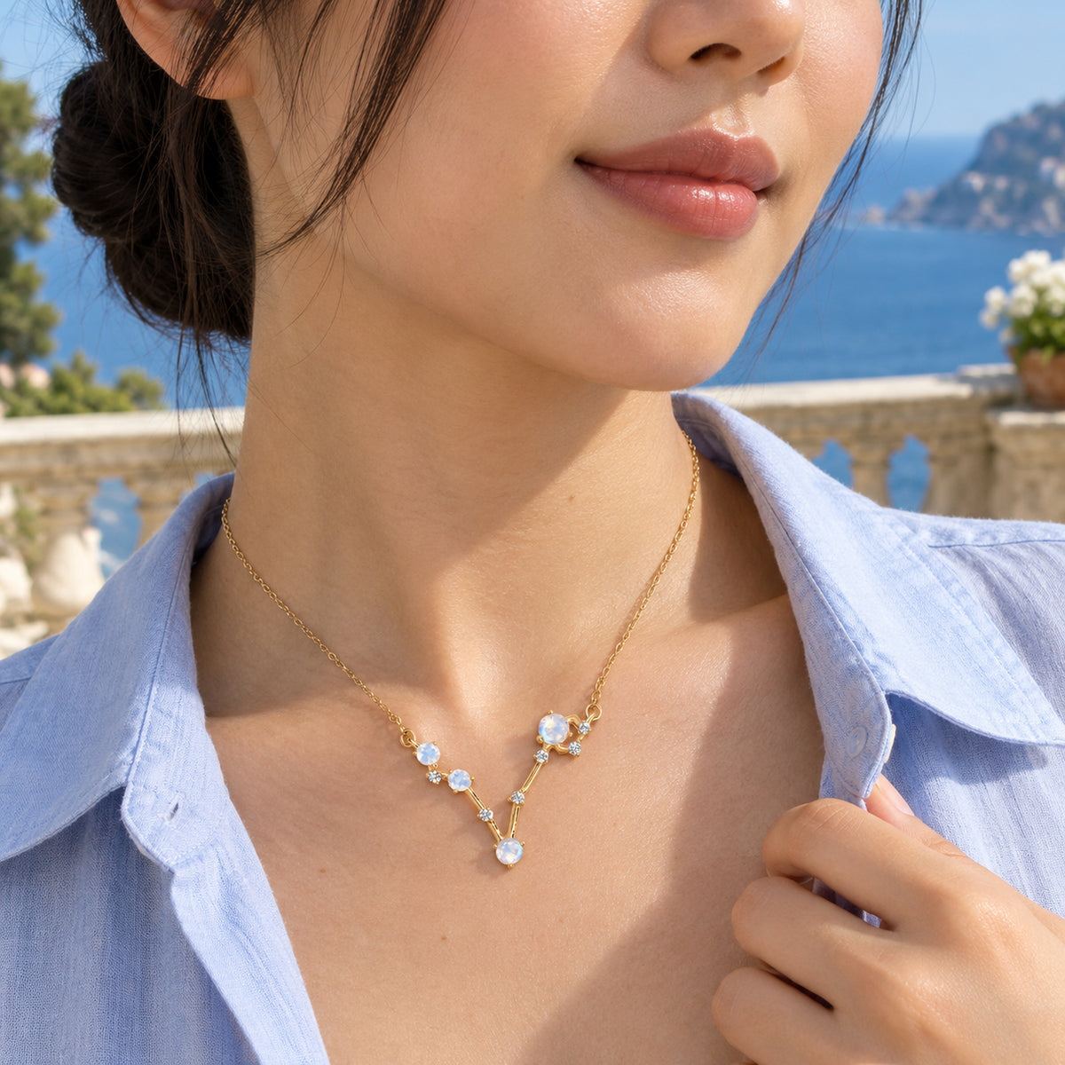 A woman with her hair in a low bun wears the gold gemstone necklace with a pale blue shirt on a seaside terrace.