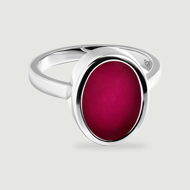 A silver ring with a deep pink oval stone rests on a light gray surface with a soft shadow.