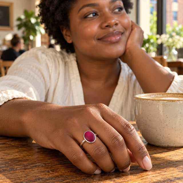 A silver ring with a pink oval stone is worn on a hand resting on a wooden cafe table beside a ceramic cup.