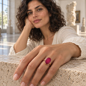 An oval pink stone set in rose gold is shown on a hand resting on a textured stone ledge in a bright gallery.