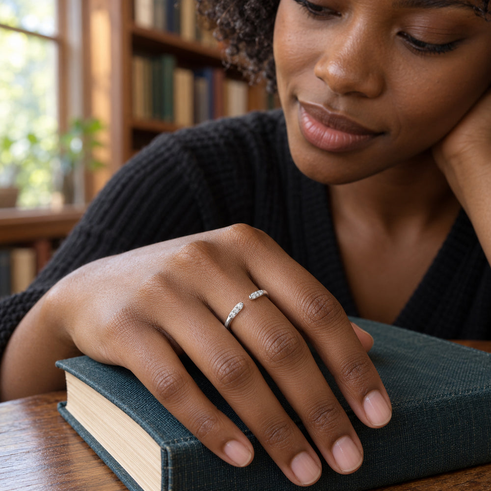 A silver open ring is worn on a hand resting on a clothbound book beside a library window.