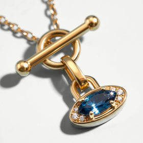 An angled close-up shows the yellow gold pendant rim, deep blue gemstone and polished bar clasp on a light gray surface.