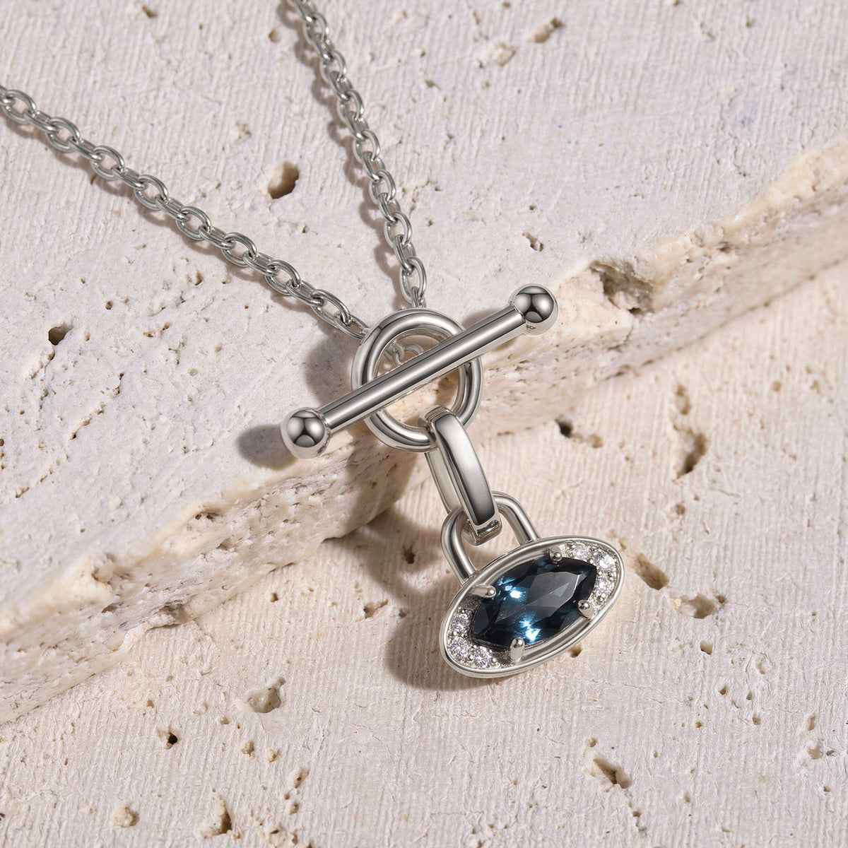 The silver necklace rests diagonally across pale textured stone with its blue gemstone pendant below the circular clasp.