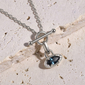 The silver necklace rests diagonally across pale textured stone with its blue gemstone pendant below the circular clasp.