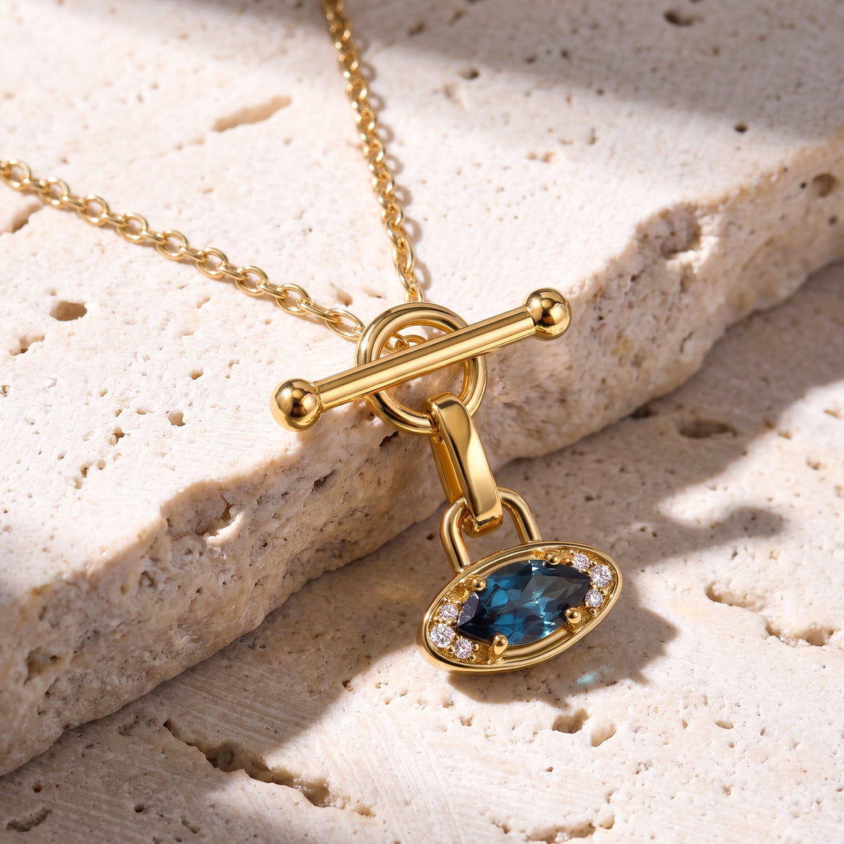 The yellow gold necklace rests diagonally across pale textured stone with its blue gemstone pendant below the circular clasp.