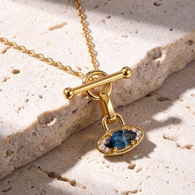 The yellow gold necklace rests diagonally across pale textured stone with its blue gemstone pendant below the circular clasp.