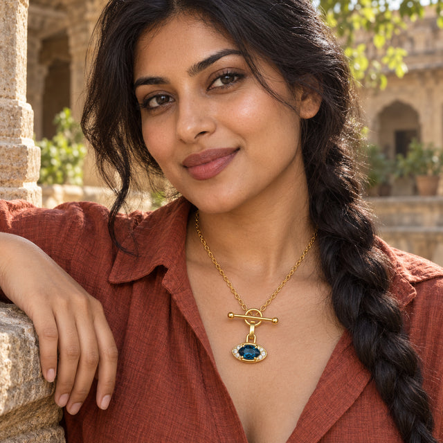 A woman in a rust-colored shirt wears the yellow gold necklace with a blue gemstone pendant in a sunlit stone courtyard.