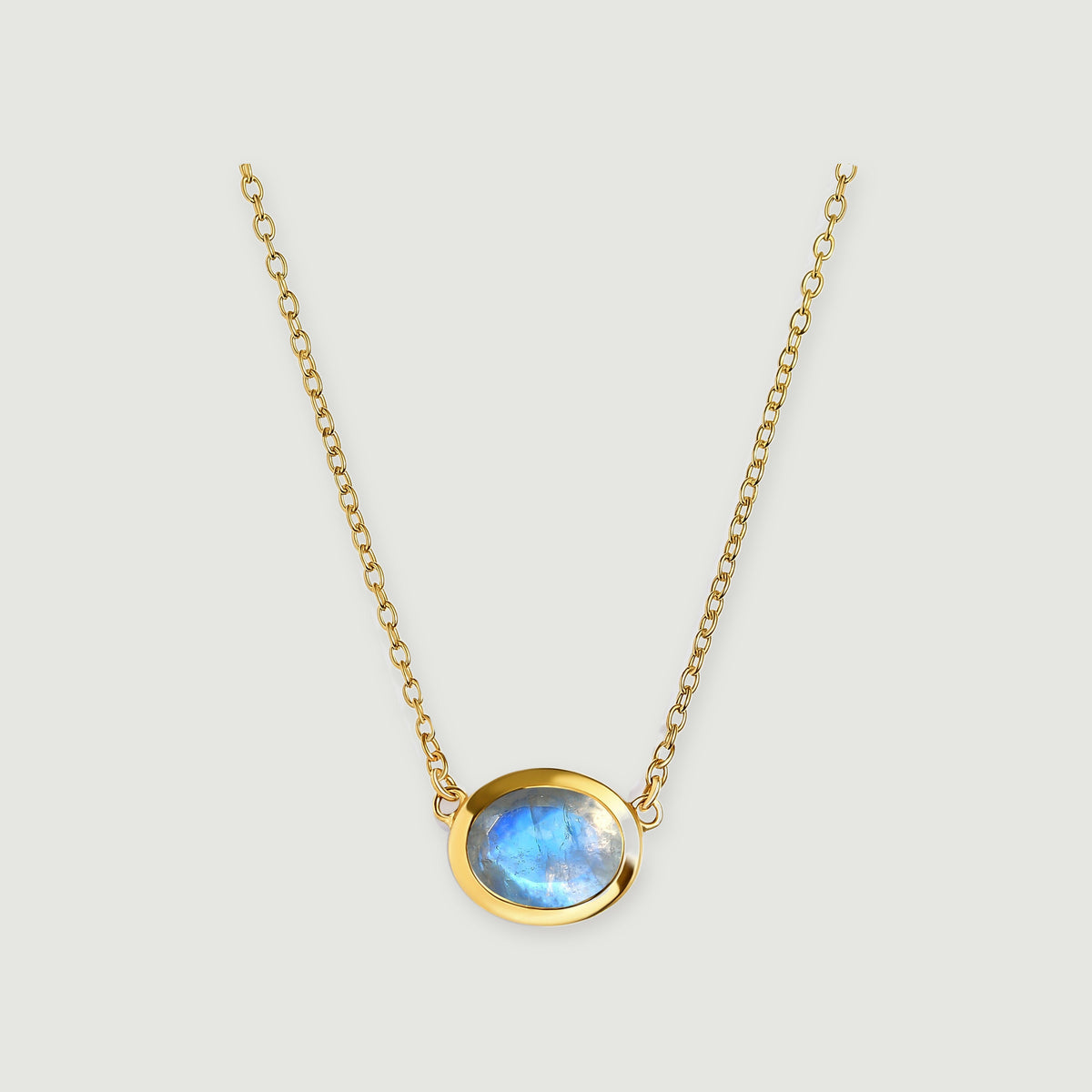 A round blue-flashing moonstone sits in a yellow gold bezel between two fine chains on a pale gray background.