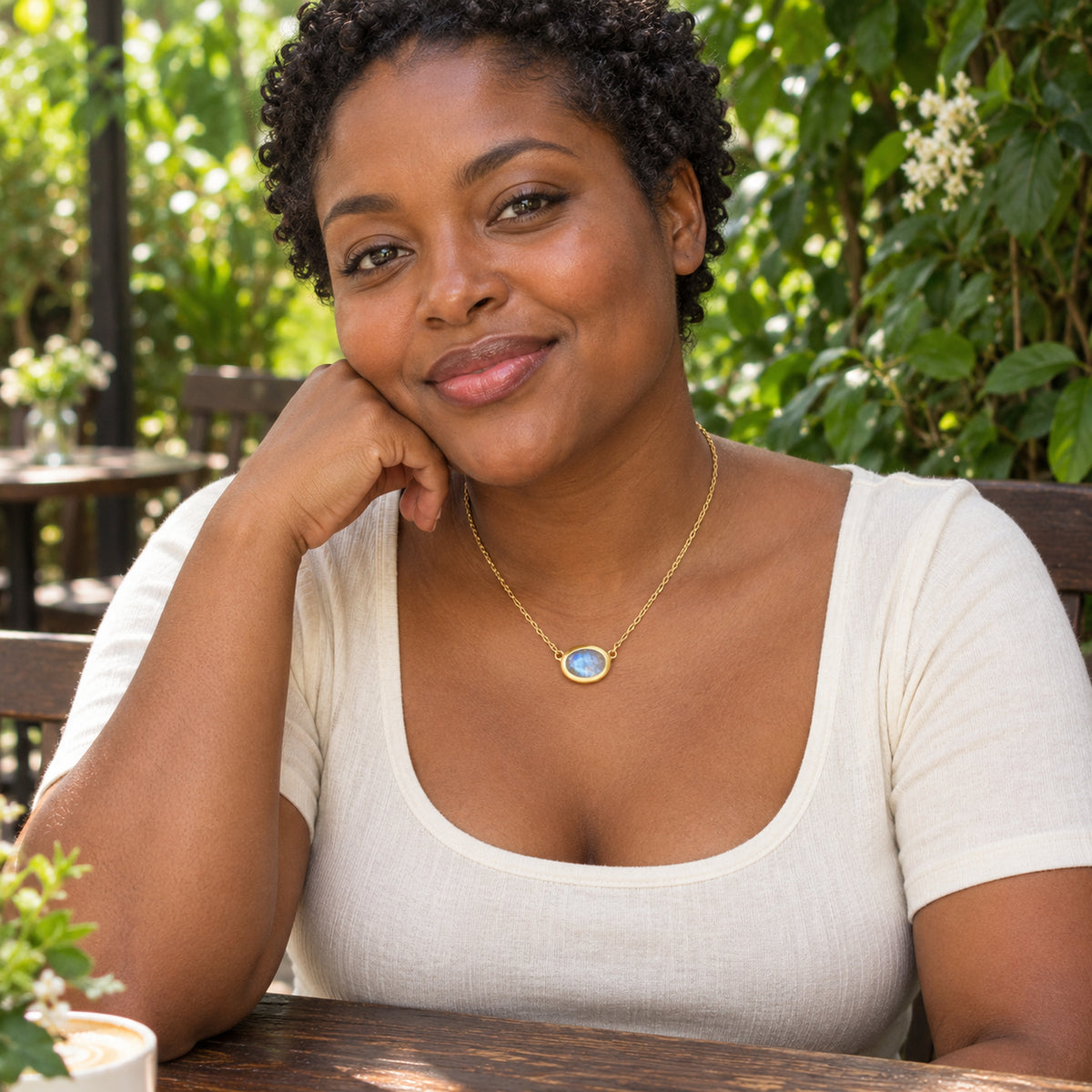 A woman with short curls wears a yellow gold moonstone necklace with an ivory top at a leafy outdoor cafe.