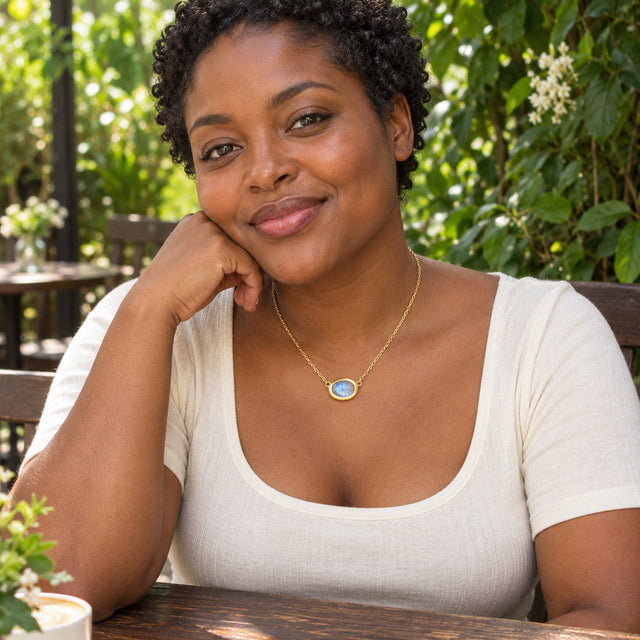 A woman with short curls wears a yellow gold moonstone necklace with an ivory top at a leafy outdoor cafe.