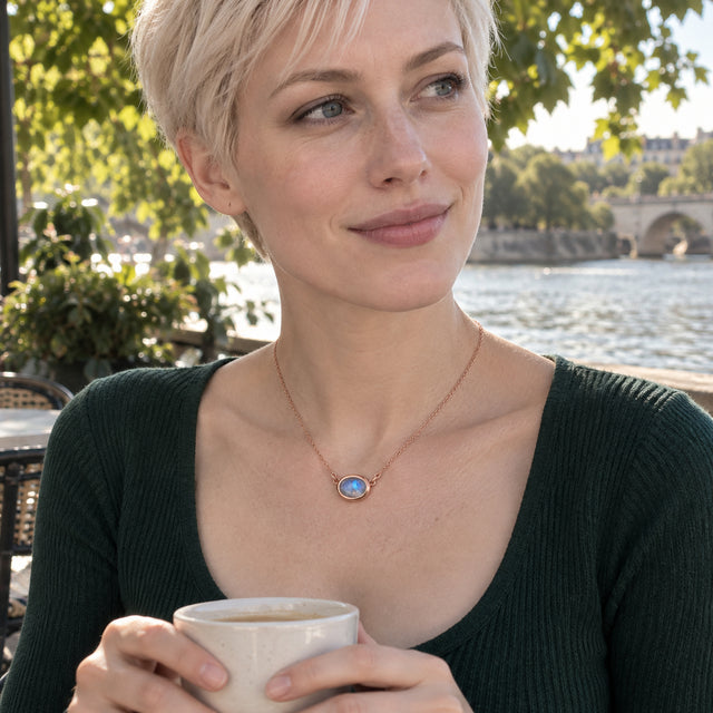 A woman with short blonde hair wears a rose gold moonstone necklace and a dark green top while holding a cup beside a river.